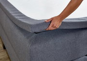 Nahaufnahme eines grauen Boxspringbetts mit einer Hand, die die Matratze anhebt, seitliche Perspektive