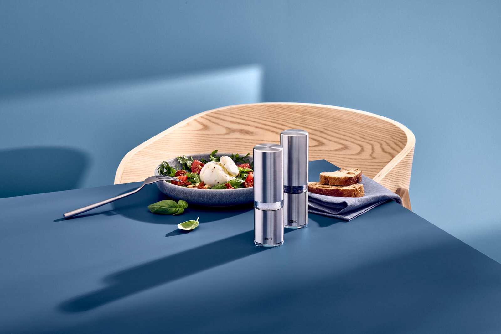 Zwei schlanke Edelstahl-Salz- und Pfeffermühlen mit transparentem Sichtfenster auf blauem Tisch vor Holzstuhl, aufgenommen in leichter Seitenansicht von vorn daneben Schale Salat, Brot und Serviette.