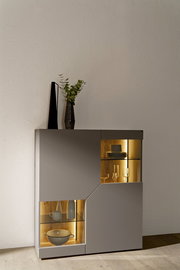 Moderne graue Vitrine/Sideboard mit integrierter warmweißer LED-Beleuchtung und Glasböden, Holzrückwand und Geschirr im Inneren, mit Vase und Deko obenauf, fotografiert in frontaler leicht seitlicher Perspektive.