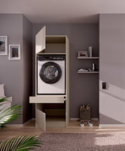 Frontaanzicht op een slanke, beige wasmachinekast met open deuren en uittrekbare plank, met een voorlader wasmachine erin, in een moderne wasruimte met grijze muur en zwevende planken.