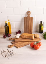 Holzgriffbrett in einer Küche, umgeben von Olivenölflaschen, Tomaten, Knoblauch und Brot. Perspektive von vorne.