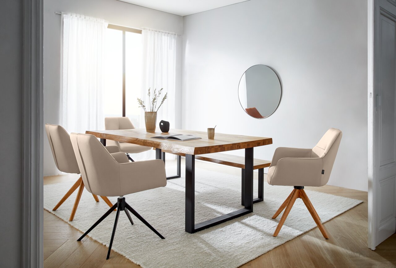 Novel Armlehnstuhl MEDUSA Moderner Essbereich mit einem großen Holztisch und mehreren beigen Armlehnstühlen auf einem hellen Teppich, seitliche Perspektive.