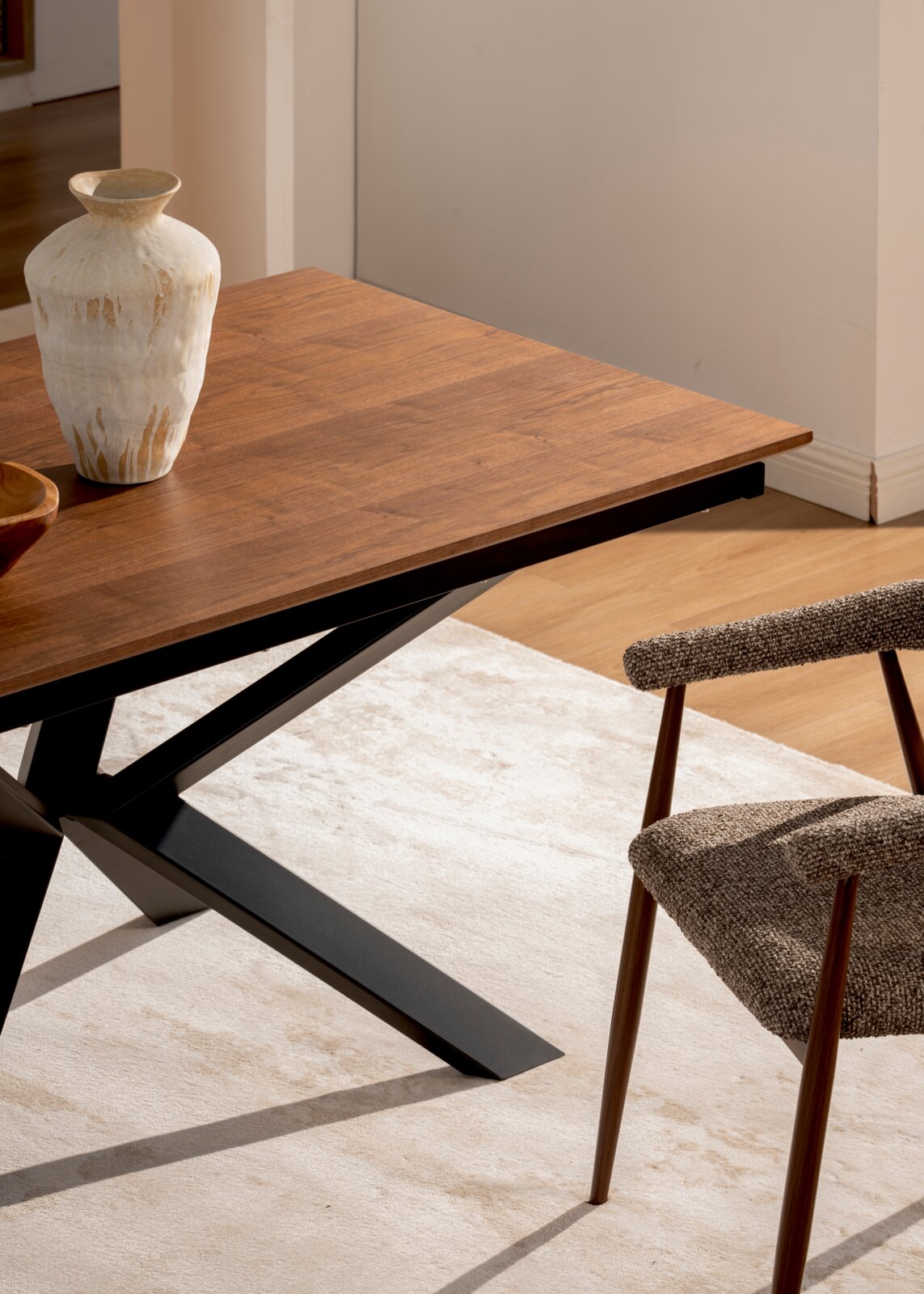 Rechthoekige houten eettafel met warmbruin blad en zwart metalen kruispoot, met vaas op het tafelblad, gefotografeerd in schuin bovenaanzicht vanuit de zijkant.
