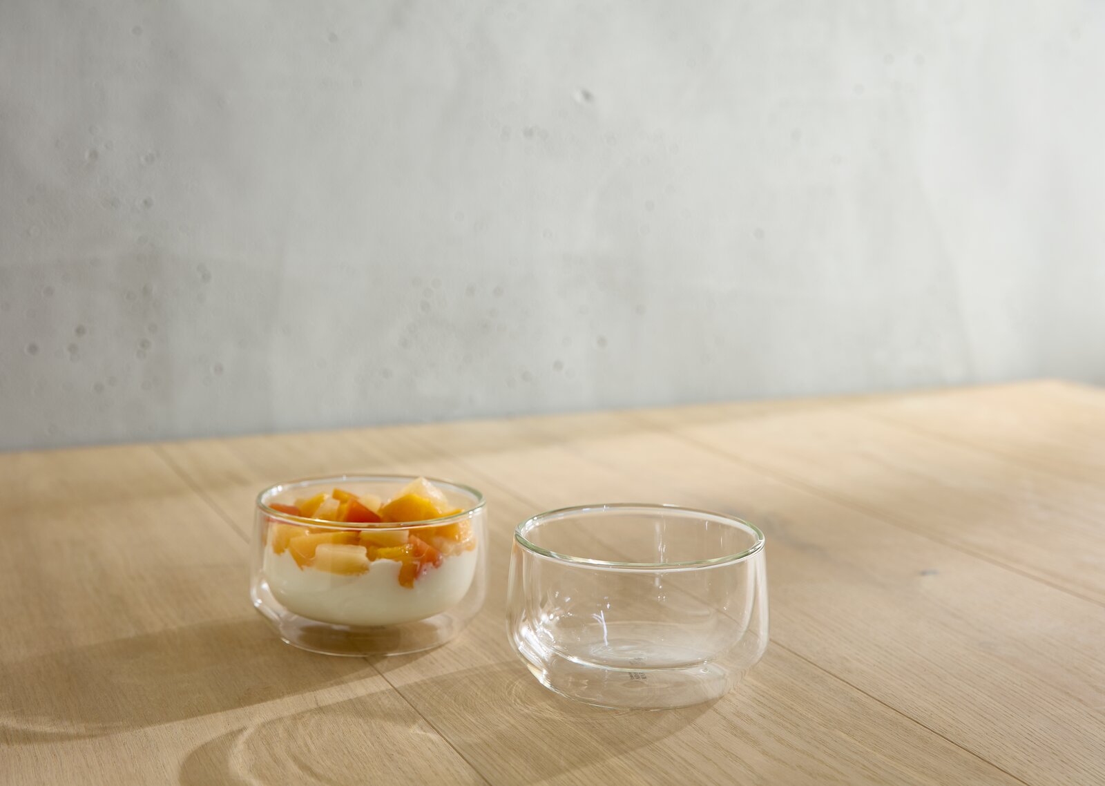 Zwei transparente Glasschalen auf einem Holztisch, eine gefüllt mit Joghurt und Obst, aus seitlicher Perspektive fotografiert.