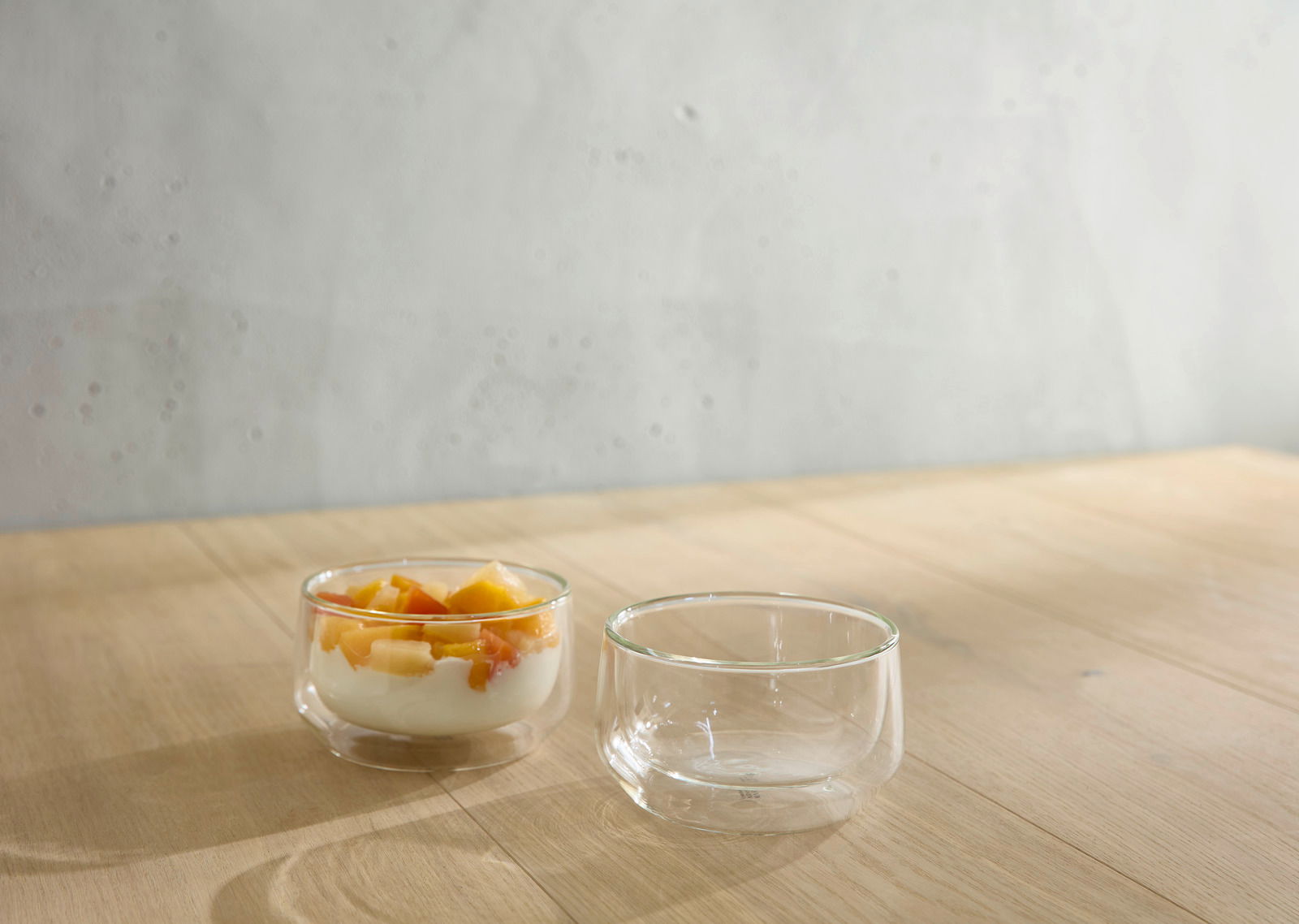 Zwei transparente Glasschalen auf einem Holztisch, eine gefüllt mit Joghurt und Obst, aus seitlicher Perspektive fotografiert.
