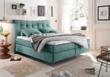 Boxspringbett in einem modernen Schlafzimmer, seitliche Perspektive, mit türkisfarbenem Kopfteil und Bettwäsche, umgeben von stilvoller Dekoration und großen Fenstern.