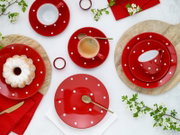 Draufsicht auf ein rot-weiß gepunktetes Kaffeeservice mit Tassen, Untertassen und Tellern, arrangiert mit Espresso, Kuchen, Löffeln, roten Servietten und Frühlingsblüten auf hellem Hintergrund