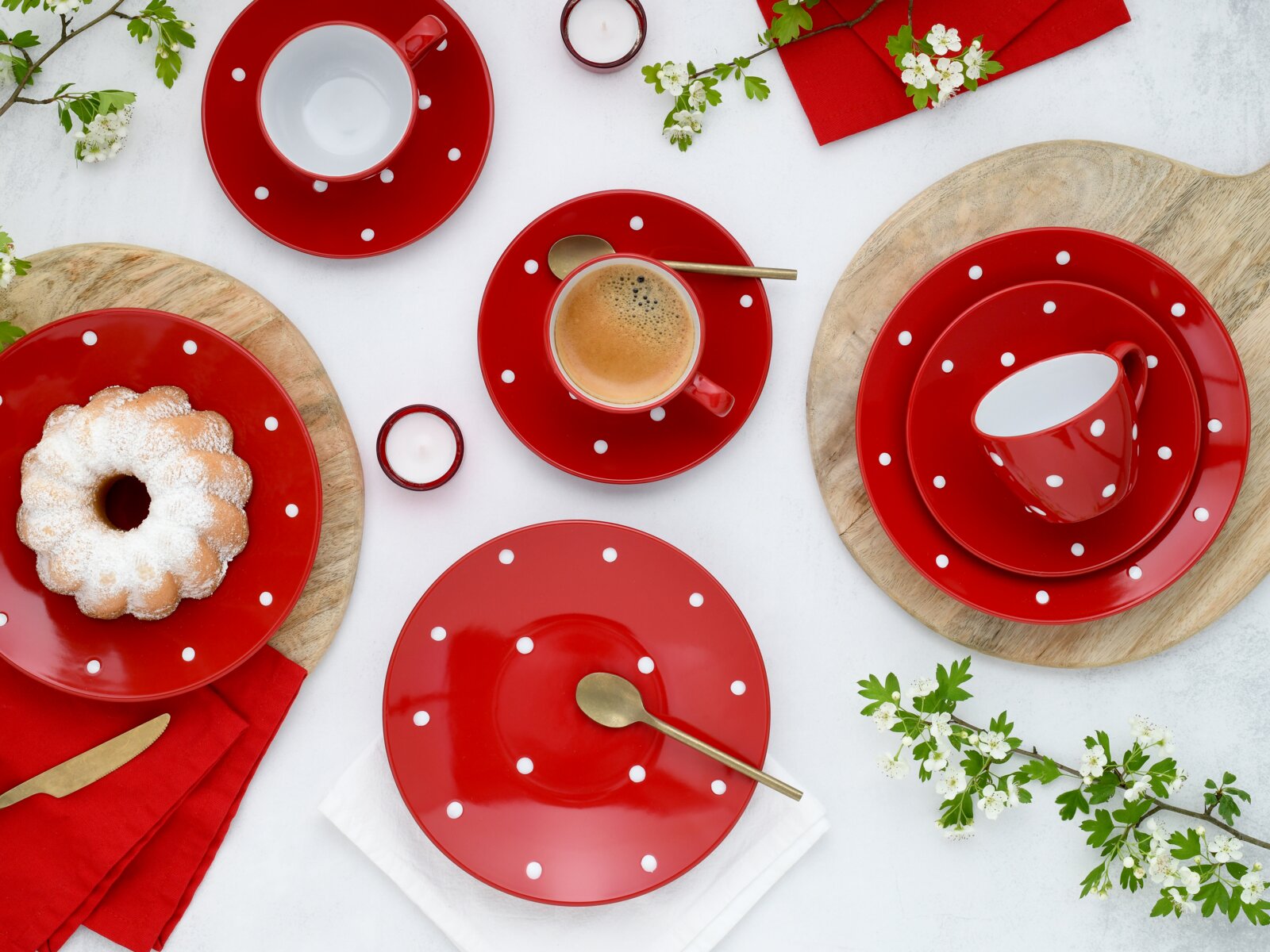 Draufsicht auf ein rot-weiß gepunktetes Kaffeeservice mit Tassen, Untertassen und Tellern, arrangiert mit Espresso, Kuchen, Löffeln, roten Servietten und Frühlingsblüten auf hellem Hintergrund