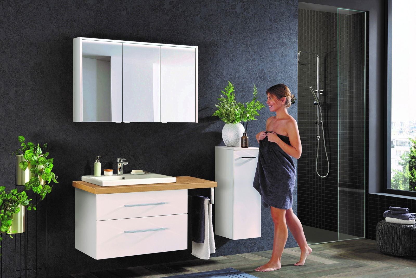 Moderne Badkombination mit weißem Waschtisch, Spiegelschrank und seitlichem Schrank, fotografiert aus einer frontalen Perspektive. Eine Frau steht daneben, umgeben von Pflanzen und einer Dusche im Hintergrund.