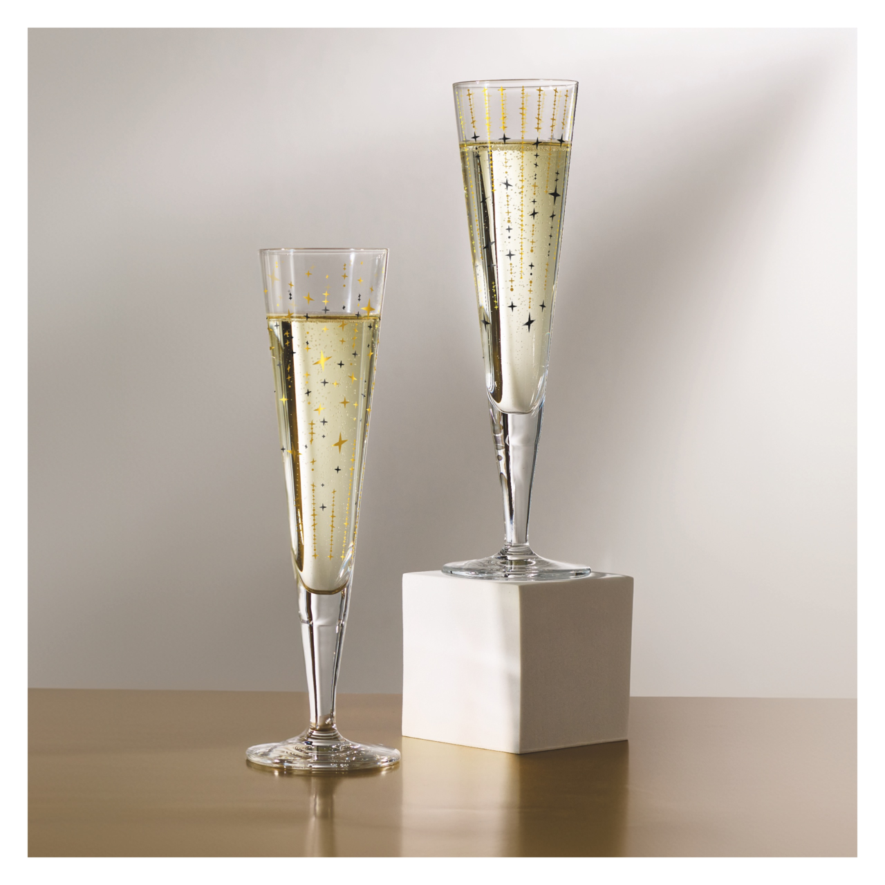 Ritzenhoff Champusglas 2er-Set GOLDNACHT Zwei elegante Champagnergläser mit goldenen Sternenmotiven, eines auf einem weißen Sockel, aus seitlicher Perspektive fotografiert.