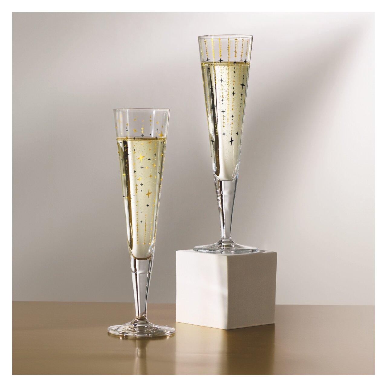 Zwei elegante Champagnergläser mit goldenen Sternenmotiven, eines auf einem weißen Sockel, aus seitlicher Perspektive fotografiert.