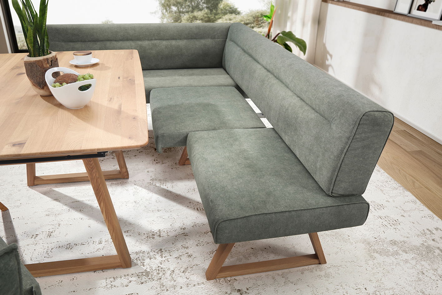 Eckbank in modernem Design mit grauem Stoffbezug und Holzbeinen, neben einem Esstisch aus Holz, aus der Perspektive von schräg oben fotografiert.