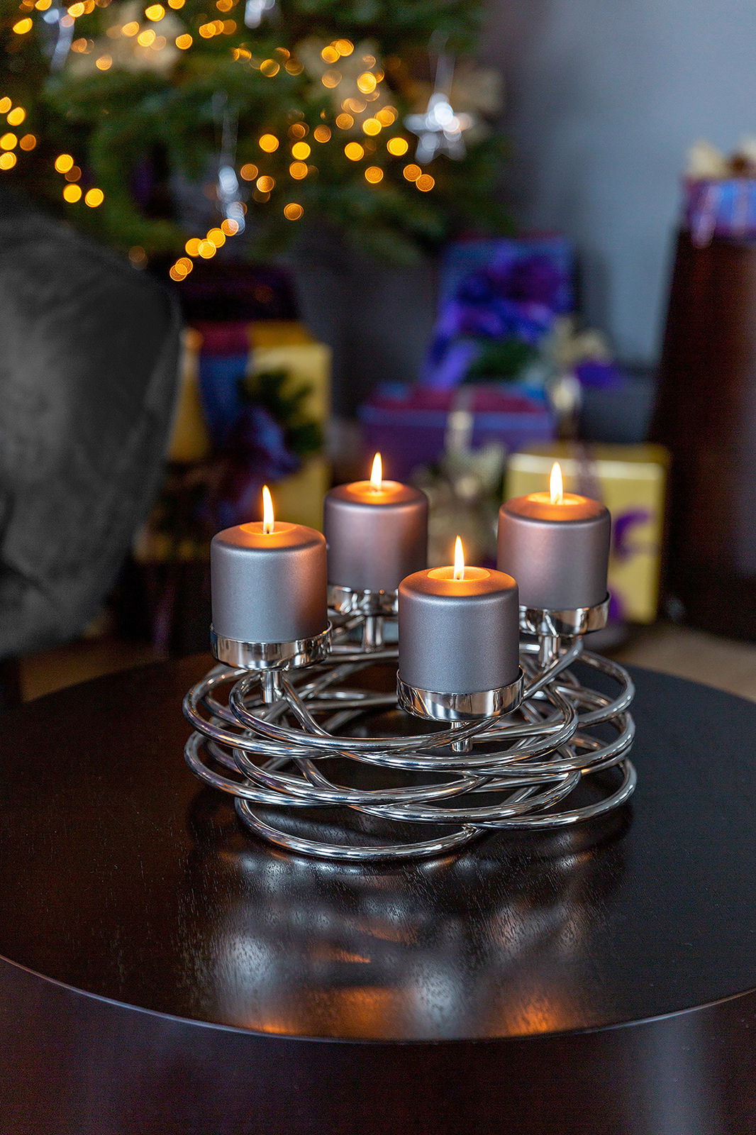Fink Dekokranz Duplex Ø35cm 4-flg. Moderner Adventskranz mit vier brennenden Kerzen auf einem Tisch, im Hintergrund ein geschmückter Weihnachtsbaum. Perspektive: seitlich.
