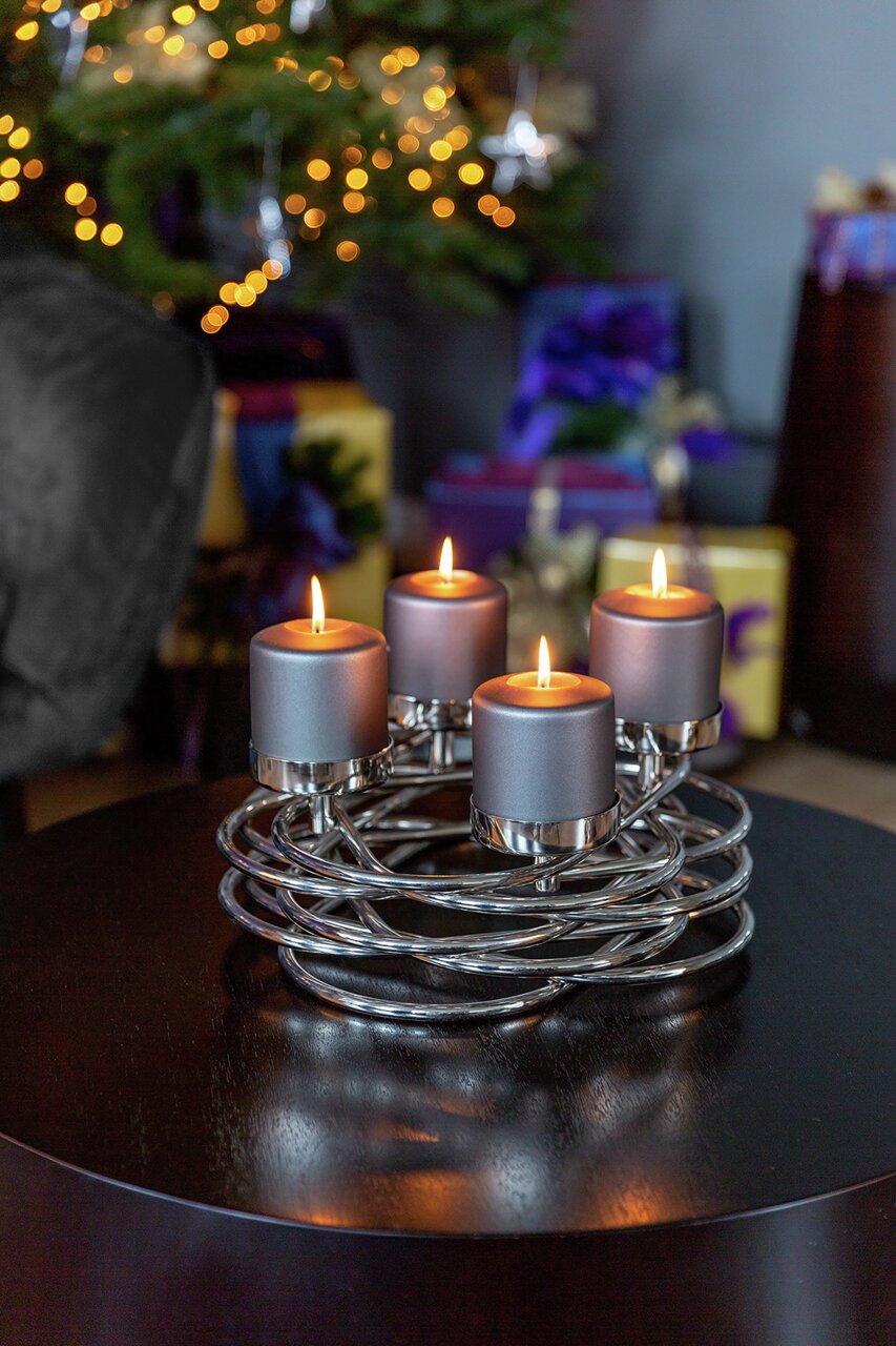 Moderner Adventskranz mit vier brennenden Kerzen auf einem Tisch, im Hintergrund ein geschmückter Weihnachtsbaum. Perspektive: seitlich.