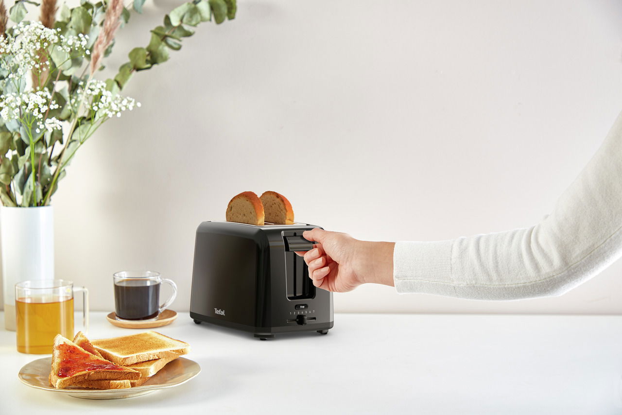 Schwarzer Toaster Vita mit zwei Scheiben Brot, seitliche Perspektive, Hand betätigt den Hebel, im Hintergrund ein gedeckter Tisch mit Tee, Kaffee und Toast.