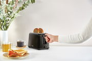 Schwarzer Toaster Vita mit zwei Scheiben Brot, seitliche Perspektive, Hand betätigt den Hebel, im Hintergrund ein gedeckter Tisch mit Tee, Kaffee und Toast.
