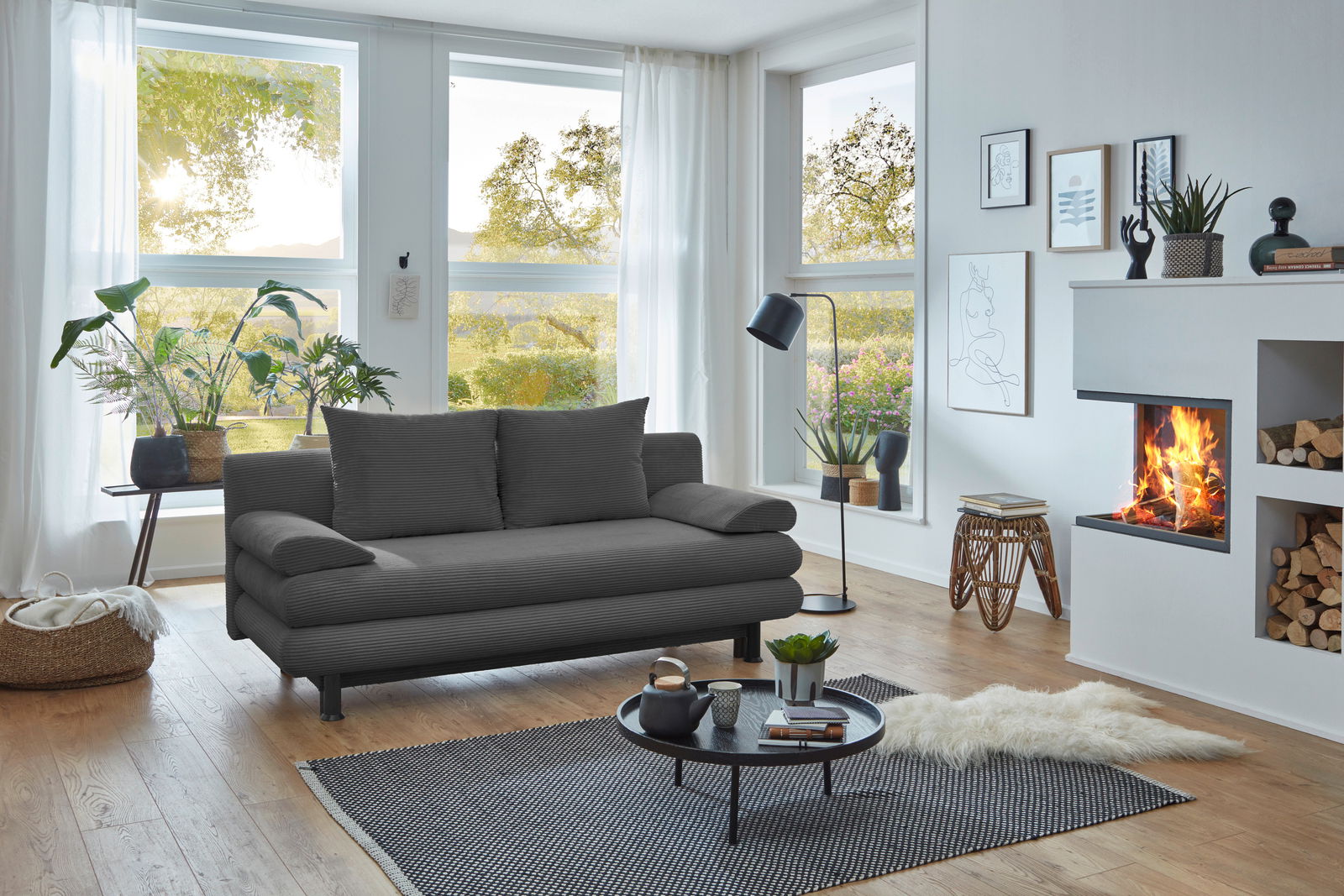 Graues Funktionssofa in modernem Wohnzimmer mit großen Fenstern, Pflanzen und Kamin, seitliche Perspektive.