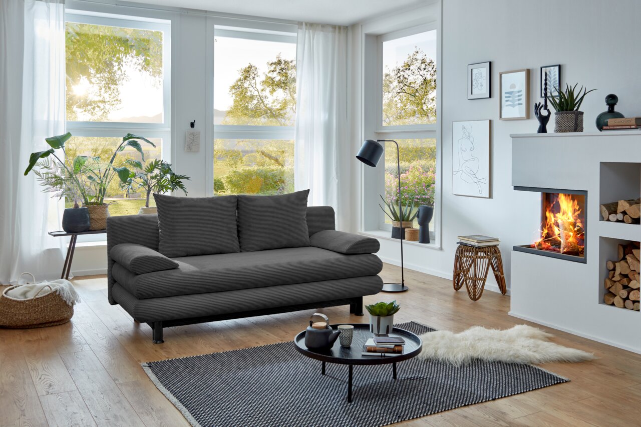 Graues Funktionssofa in modernem Wohnzimmer mit großen Fenstern, Pflanzen und Kamin, seitliche Perspektive.