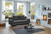 Graues Funktionssofa in modernem Wohnzimmer mit großen Fenstern, Pflanzen und Kamin, seitliche Perspektive.
