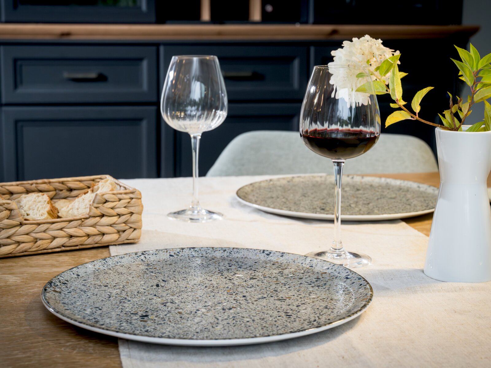 Gedeckter Esstisch mit zwei gesprenkelten Keramiktellern, zwei Weingläsern (eins mit Rotwein), Brotkorb und weißer Vase mit Blüten, frontal aus leichter Tischhöhe aufgenommen