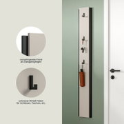 Schmale Wandgarderobe/Schlüsselboard in Weiß mit schwarzem Rahmen und schwarzen Metallhaken, an der Wand montiert mit Schlüsselbund und Tasche; frontale Ansicht mit leichter Seitenperspektive.