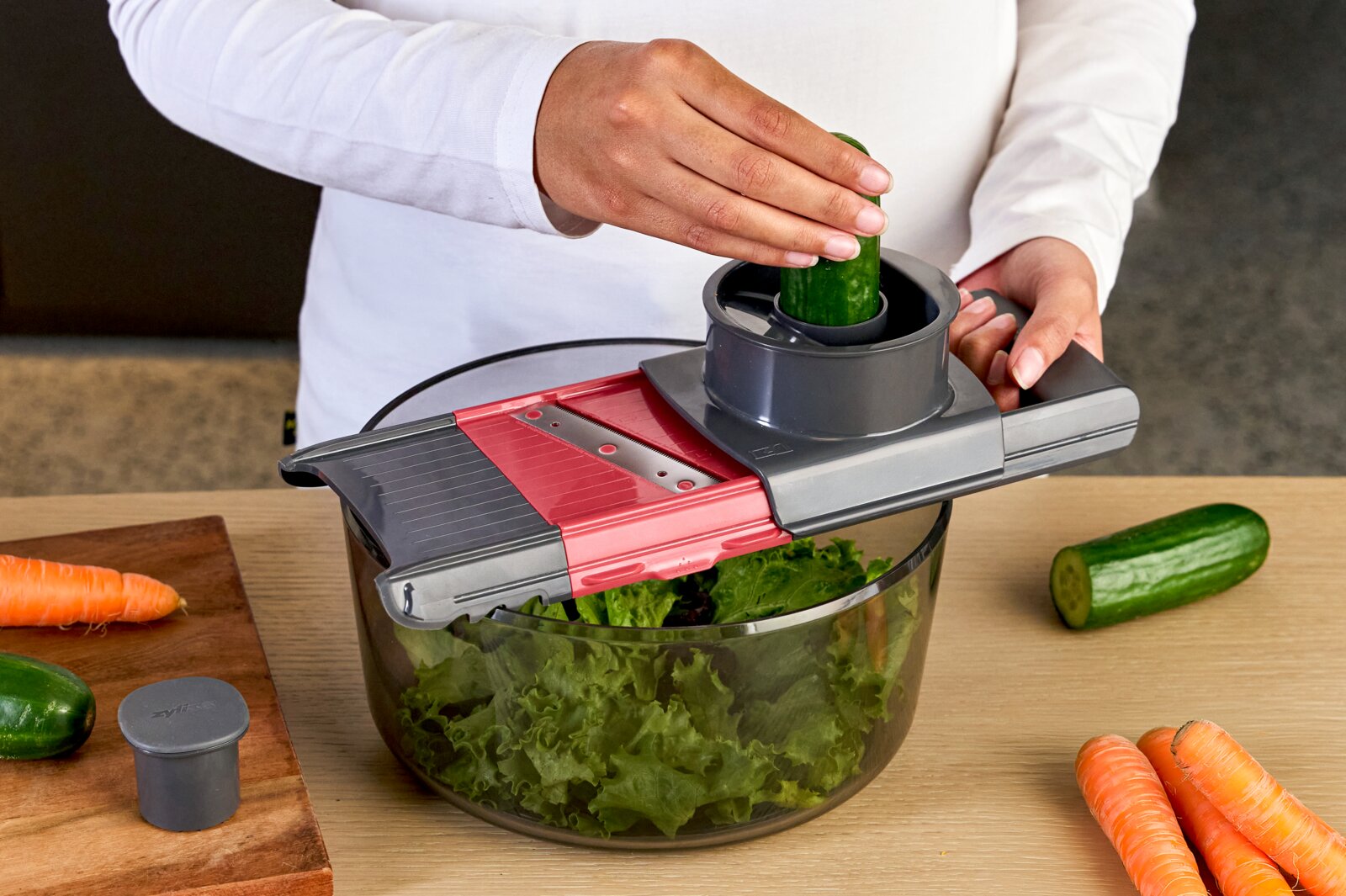 Schüssel mit Gemüseschneider/Mandoline in Grau-Rot und Fingerschutz, Gurke wird von oben in den Einfüllschacht gedrückt; Frontansicht leicht von oben auf einem Küchentisch mit Salat in der Schüssel und Karotten daneben.