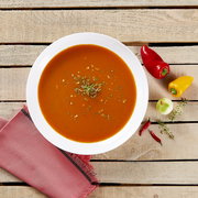Draufsicht auf eine Schüssel würzige rote Paprika-Tomatensuppe, garniert mit Kräutern, auf rustikalem Holztisch mit roten und gelben Paprika, Zwiebel und Chilischoten.
