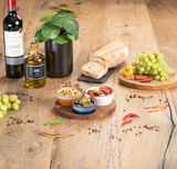 Servierbrett mit drei Schalen, gefüllt mit verschiedenen Antipasti, auf einem Holztisch mit Weinflasche, Ölflasche, Brot, Trauben und Gewürzen. Draufsicht.