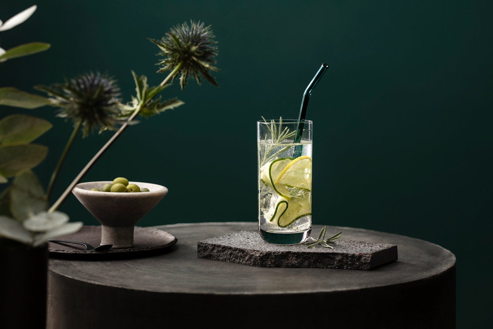 Ein Longdrinkbecher mit einem Getränk, Gurkenscheiben und einem grünen Strohhalm, auf einem dunklen Tisch mit einer Schale Oliven und dekorativen Pflanzen im Hintergrund, aus seitlicher Perspektive.