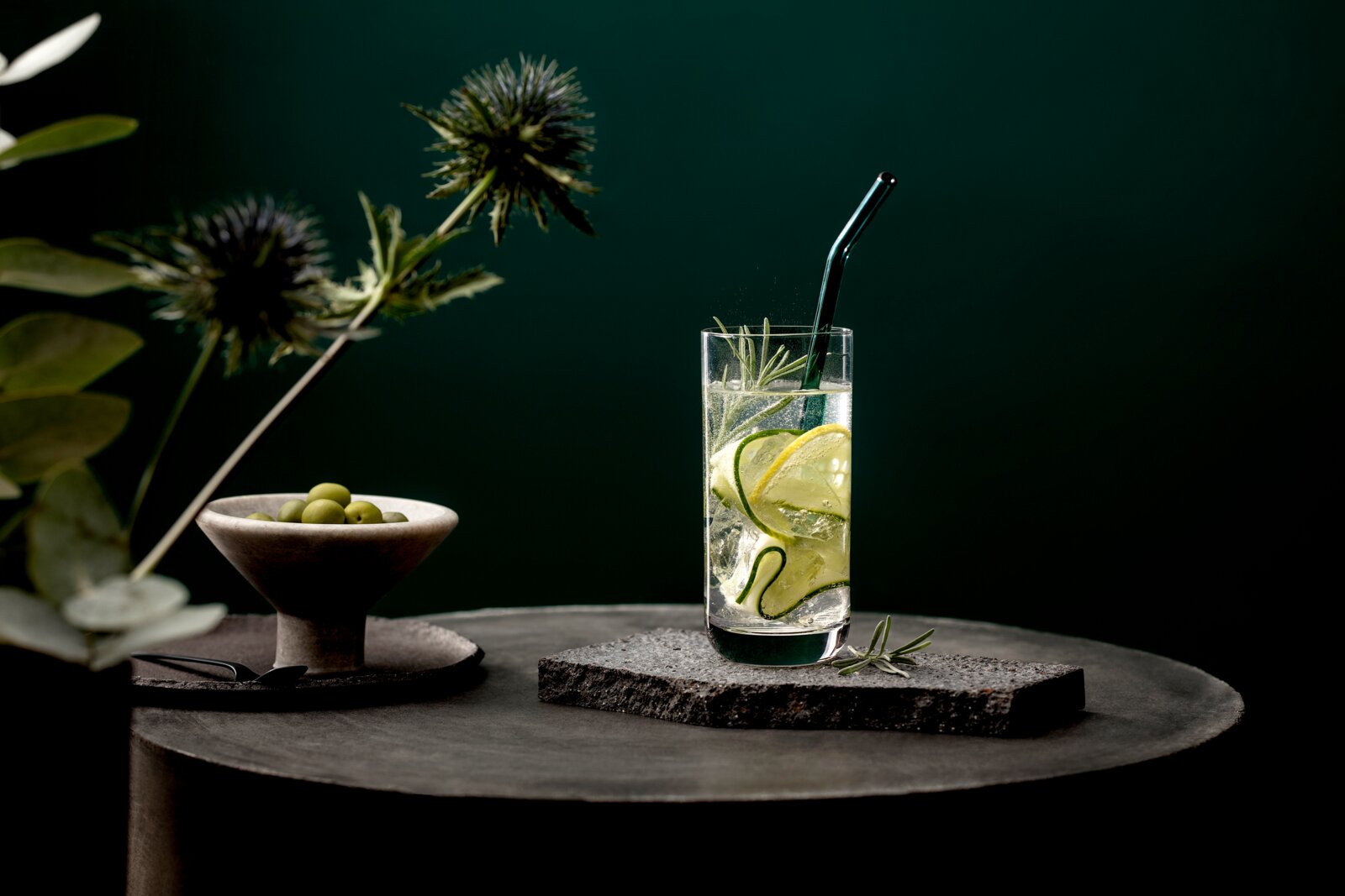 Ein Longdrinkbecher mit einem Getränk, Gurkenscheiben und einem grünen Strohhalm, auf einem dunklen Tisch mit einer Schale Oliven und dekorativen Pflanzen im Hintergrund, aus seitlicher Perspektive.