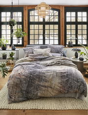 Gemütliches Schlafzimmer mit irisette Bettwäsche ZOBEL in natürlichem Design, fotografiert aus der Frontperspektive. Die Bettwäsche zeigt ein dezentes, erdiges Muster und ist auf einem stilvoll eingerichteten Bett in einem hellen Raum mit großen Fenstern