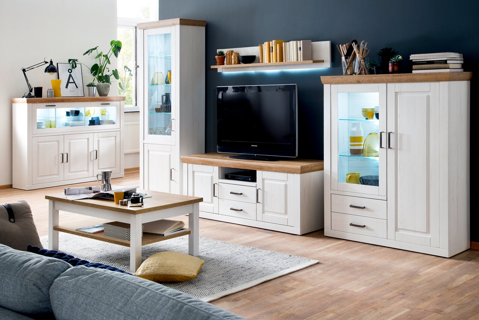 Wohnzimmermöbel-Set im Landhausstil in Weiß mit Holzoptik-Deckplatten, bestehend aus Vitrine, Sideboard, TV-Lowboard und Highboard mit Glasfront und Beleuchtung; dargestellt in Frontansicht mit leicht schräger Perspektive von links.