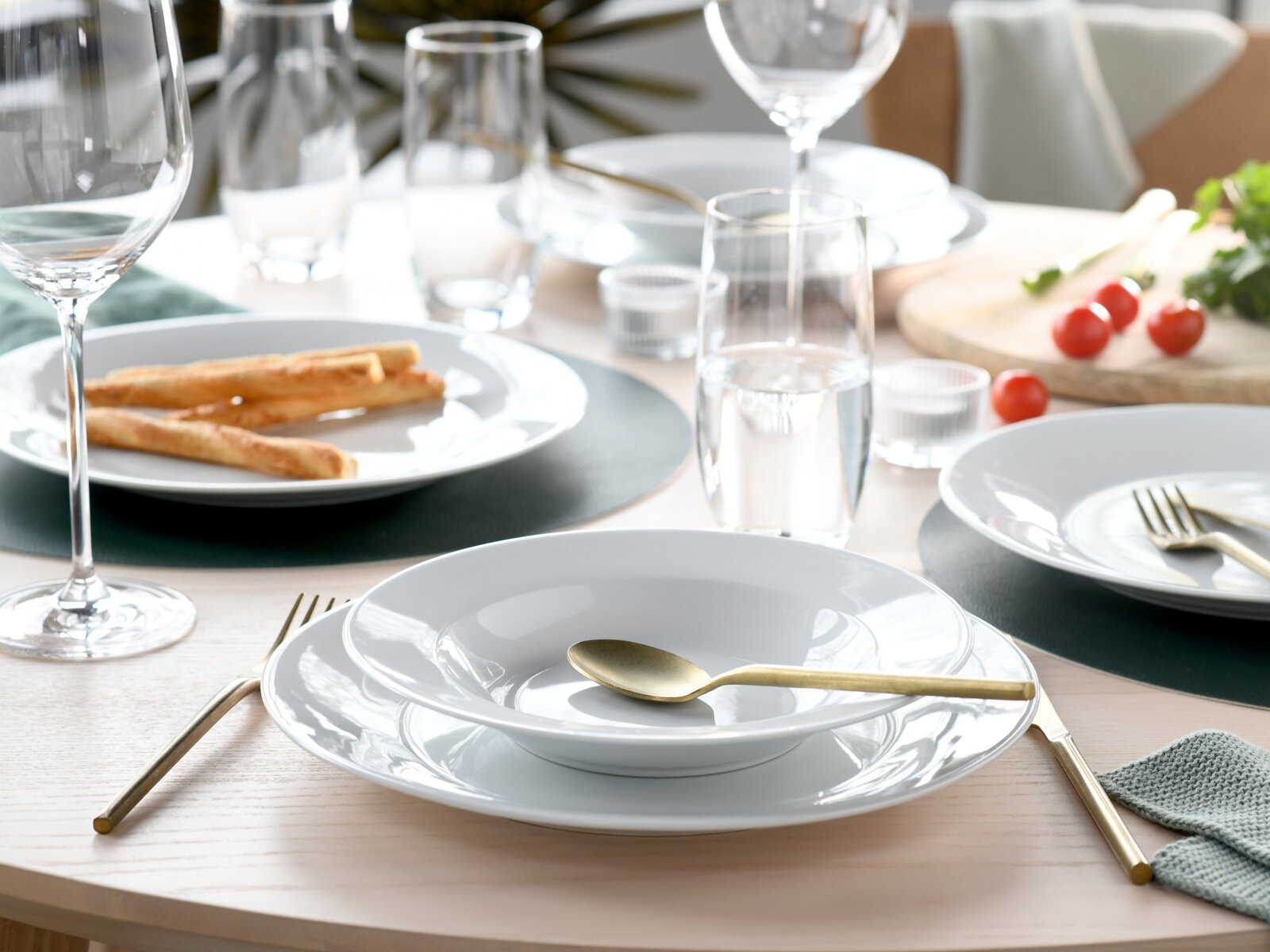Elegant gedeckter Esstisch mit weißem Geschirr, goldfarbenem Besteck und Weingläsern; Aufnahme aus leicht erhöhter, schräger Perspektive von vorne.