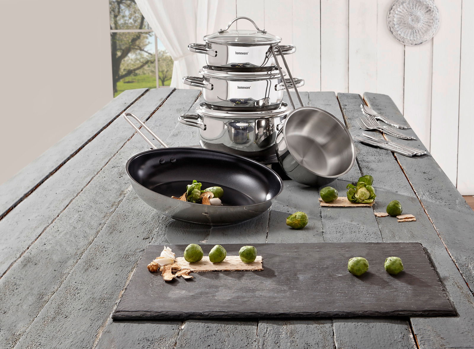 Topfset 5-tlg. aus Edelstahl, bestehend aus gestapelten Töpfen und einer Pfanne, auf einem rustikalen Holztisch mit Blick auf ein Fenster im Hintergrund.