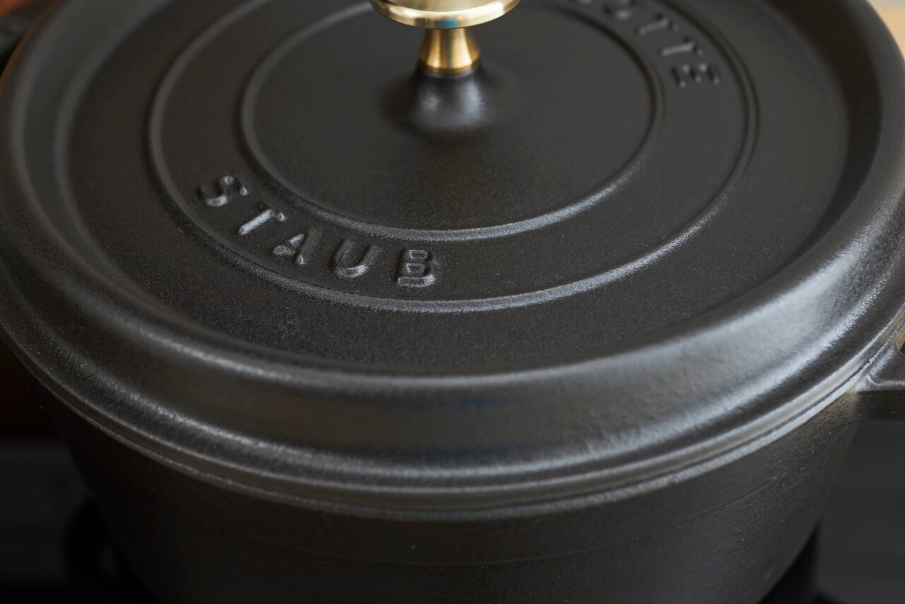 Close-up van een ronde, zwarte braadpan met deksel en goudkleurige knop, van het merk Staub, gezien van bovenaf.