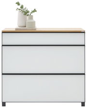Moderne ladekast met drie greeploze lades, lichtgrijze fronten en een zwart frame, evenals een houten blad, versierd met een vaas en eucalyptus; vooraanzicht op een witte achtergrond.