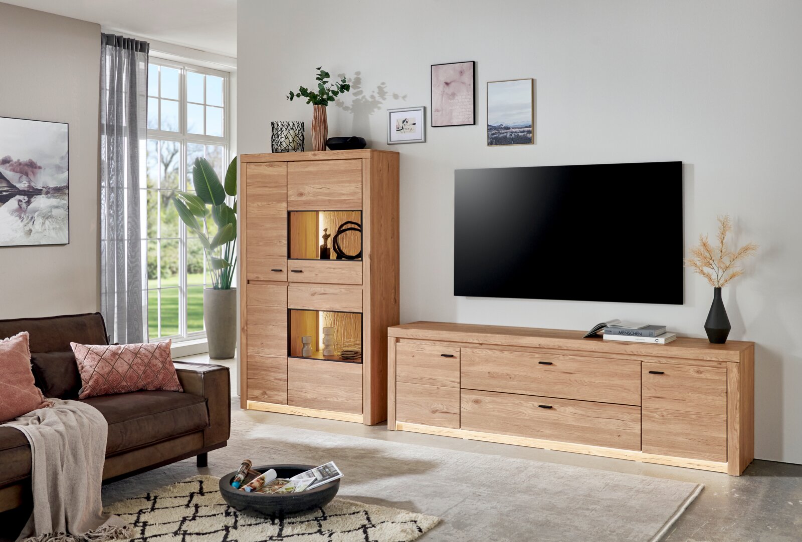 Cantus Wandkombination WIEN Modernes Wohnzimmer mit TV-Lowboard und Vitrine aus hellem Holz mit integrierter Beleuchtung, frontal aus leicht schräger Perspektive aufgenommen