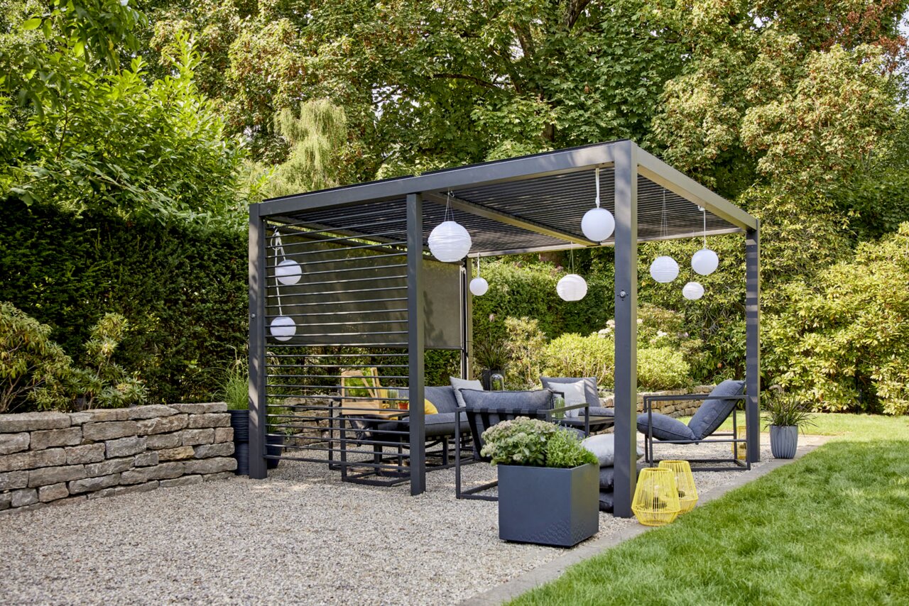 Moderne Pergola aus grauem Metall mit Lamellendach und seitlichem Sichtschutz, eingerichtet als Lounge mit Hängelaternen im grünen Garten, perspektivisch in schräger Vorderansicht von links.