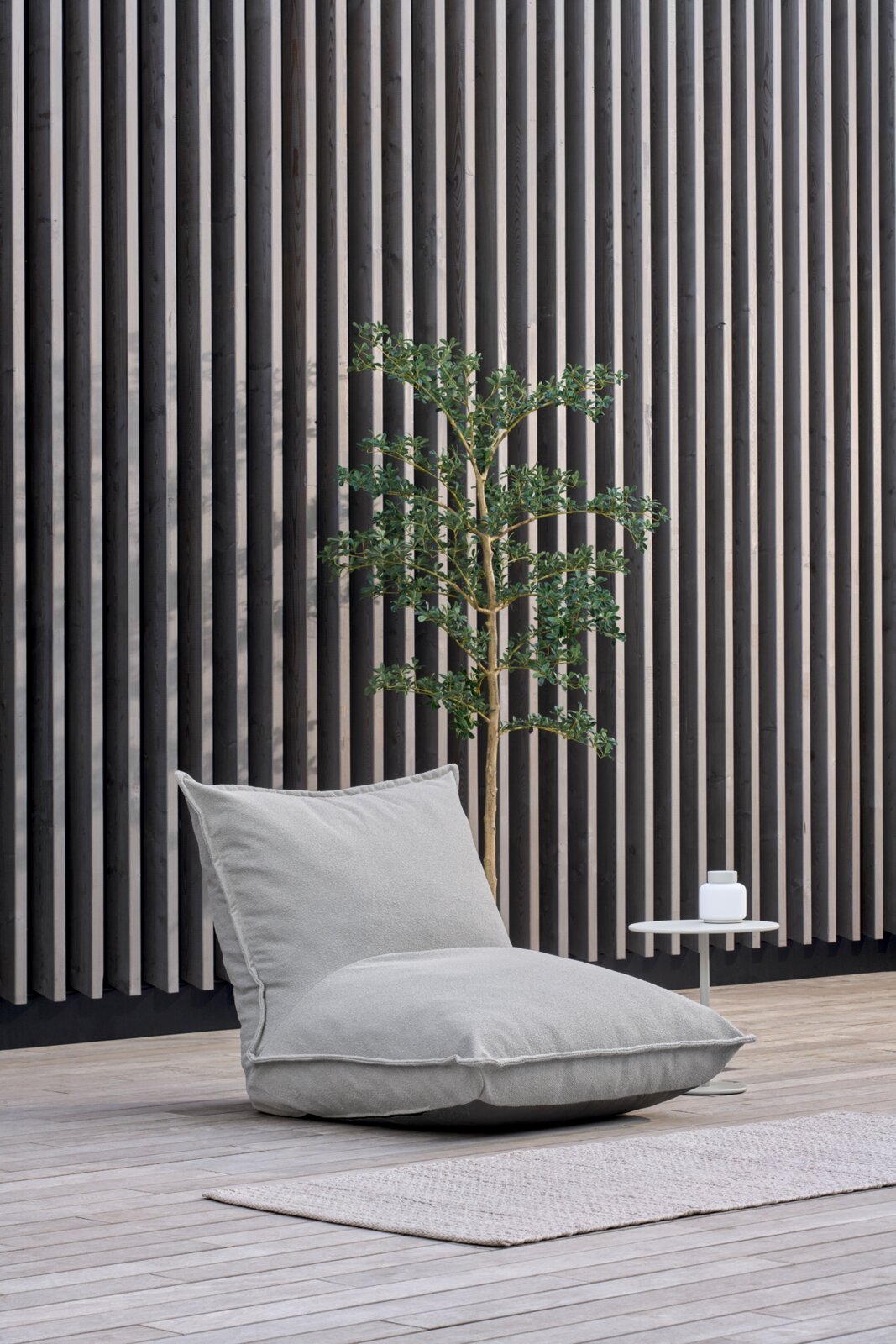 Lichtgrijze zitzak-loungestoel met rugleuning op een houten terras voor een verticale wand van houten lamellen, zijaanzicht