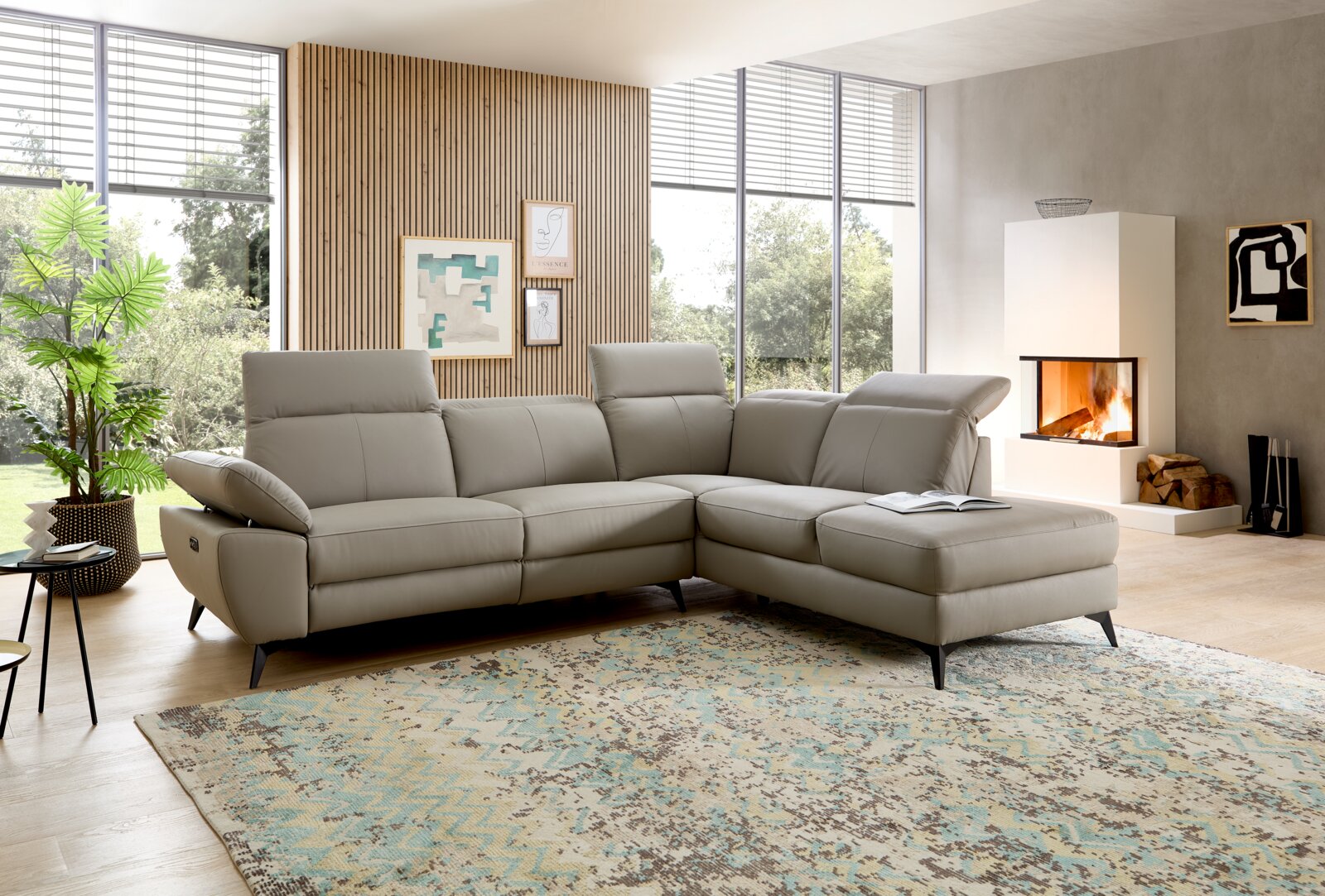 Modernes graues L-förmiges Ecksofa aus Leder/lederähnlichem Material mit verstellbaren Kopfstützen und Recamiere rechts, auf Teppich im hellen Wohnzimmer; Perspektive: frontale leicht schräge Ansicht von links.