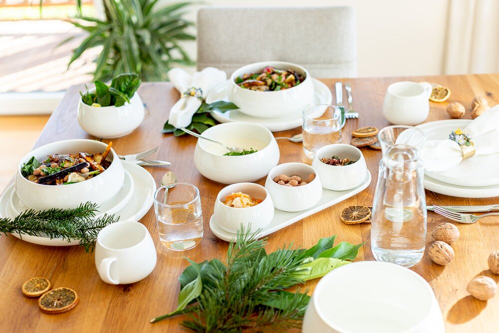 Modern gedeckter Holztisch mit weißem Geschirr-Set (Schalen, Teller, kleine Schälchen), Wasserkrug und Gläsern, dekoriert mit Tannenzweigen, Nüssen und getrockneten Orangenscheiben; Perspektive: leicht erhöhte, schräge Ansicht von vorne.