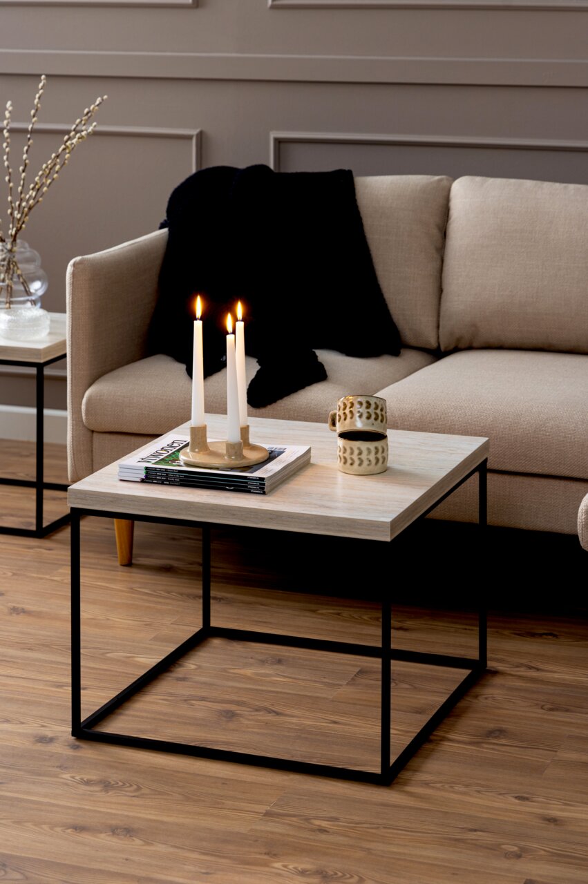 Moderner Couchtisch mit heller Holzplatte und schwarzem Metallgestell, dekoriert mit Kerzen und Tasse, seitliche Perspektive.
