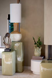 Modernes Wohnzimmer mit einem dekorativen Ornament auf einem glänzenden, grünen Sockel. Im Hintergrund eine Tischlampe mit weißem Schirm und eine Vase mit Pflanzen. Seitliche Perspektive.