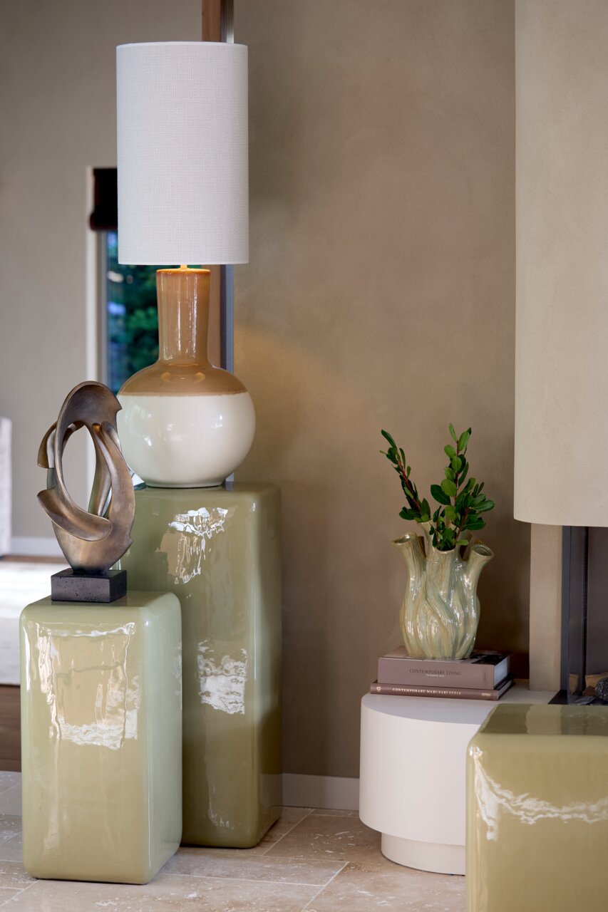 Modernes Wohnzimmer mit einem dekorativen Ornament auf einem glänzenden, grünen Sockel. Im Hintergrund eine Tischlampe mit weißem Schirm und eine Vase mit Pflanzen. Seitliche Perspektive.