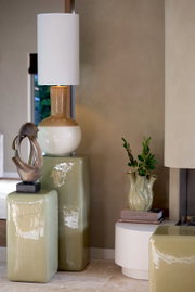 Modernes Wohnzimmer mit einem dekorativen Ornament auf einem glänzenden, grünen Sockel. Im Hintergrund eine Tischlampe mit weißem Schirm und eine Vase mit Pflanzen. Seitliche Perspektive.