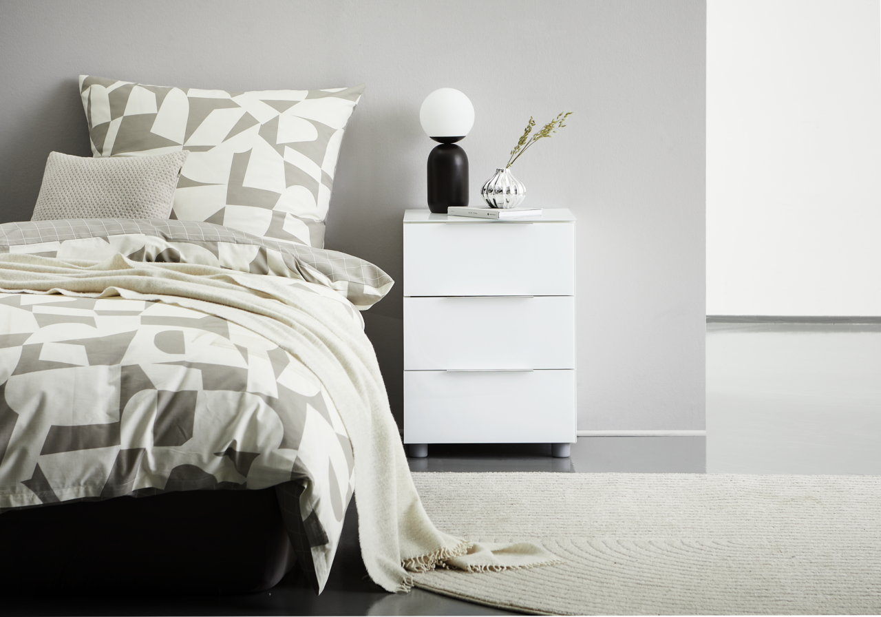 Voleo Nachtkonsole MALIN Weiße Nachtkonsole mit drei Schubladen, dekoriert mit einer Lampe und einer Vase, seitliche Perspektive