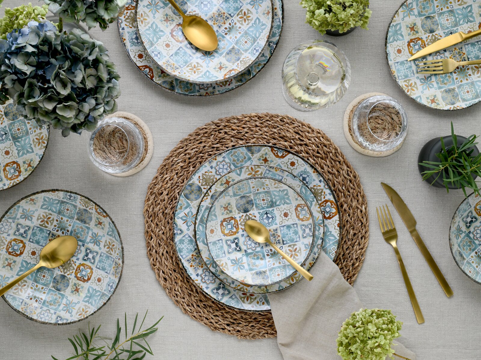 Draufsicht auf ein stilvoll gedecktes Tischarrangement mit gemusterten Keramiktellern in Blau- und Ockertönen, goldfarbenem Besteck, geflochtenen Platzsets, Gläsern und grünen Hortensien auf hellem Leinentuch.