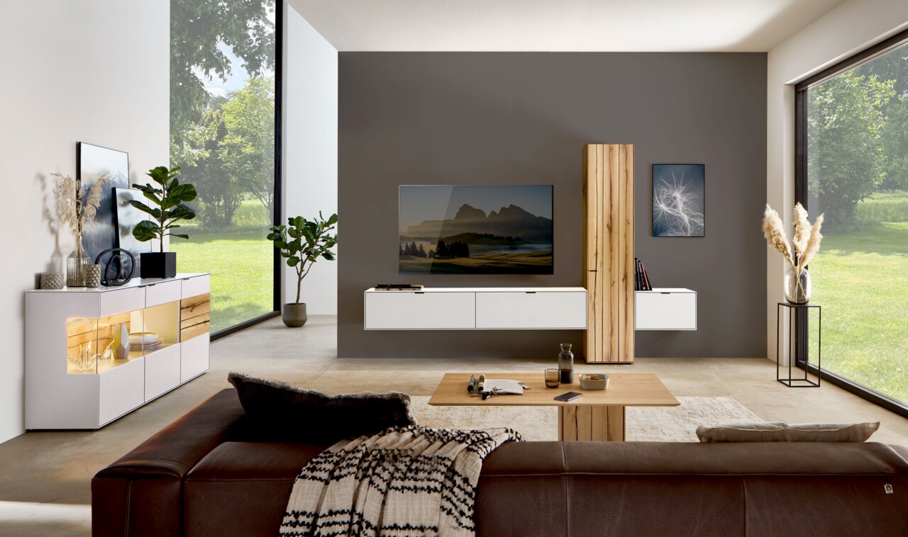 Moderne Wohnzimmer-Wandkombination in einem hellen Raum mit großen Fenstern, bestehend aus weißen und holzfarbenen Elementen, mit einem Fernseher an der Wand. Perspektive von der Seite.