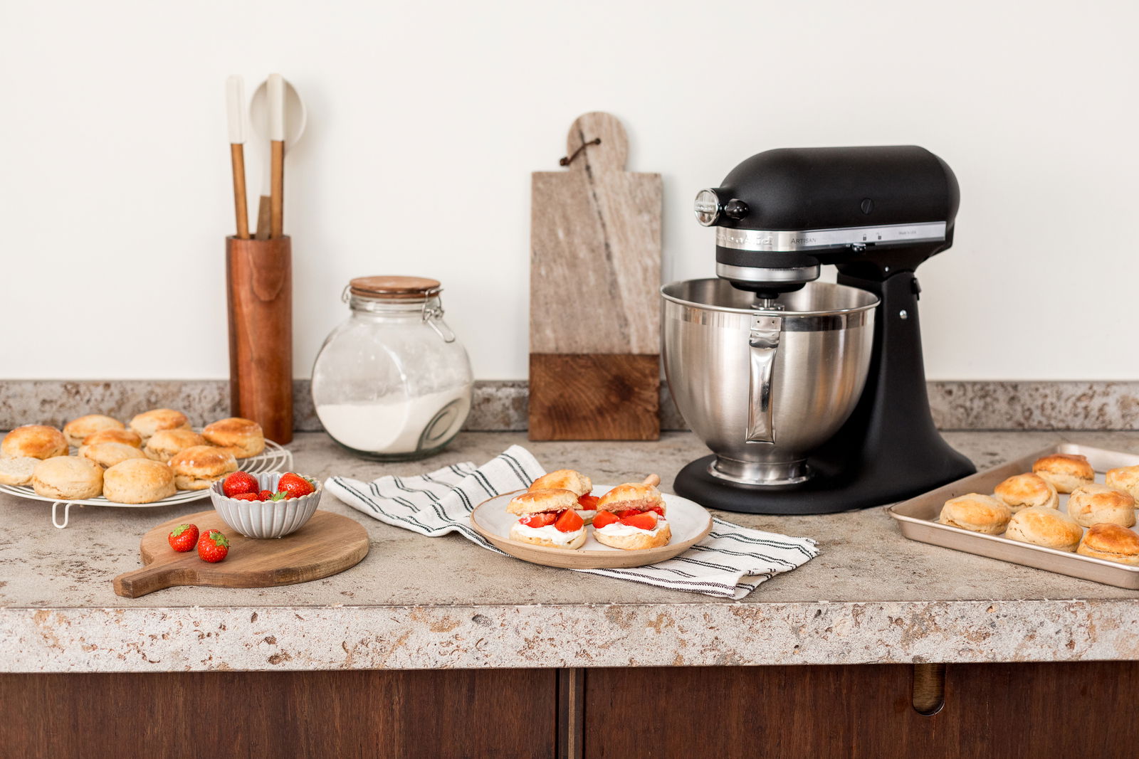 Schwarze Küchenmaschine aus Guss auf einer Küchenarbeitsplatte, umgeben von Backutensilien und frischen Brötchen, seitliche Perspektive.