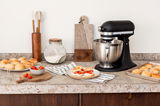 Schwarze Küchenmaschine aus Guss auf einer Küchenarbeitsplatte, umgeben von Backutensilien und frischen Brötchen, seitliche Perspektive.